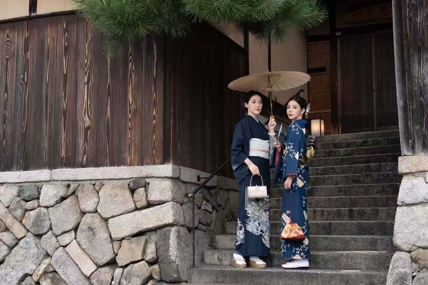 Two individuals in traditional attire on stone steps with a wooden fence and a pine tree in the background. 3