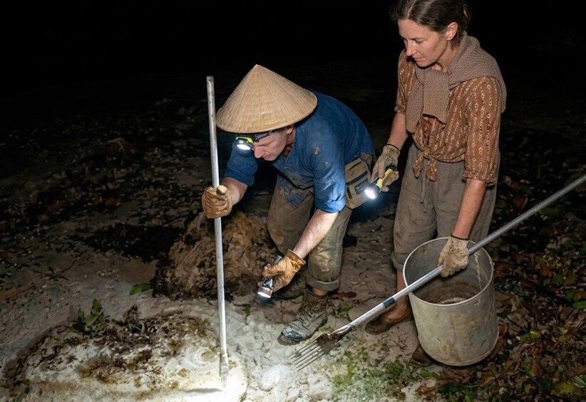 Two individuals engaged in an outdoor activity, possibly involving soil or sediment, with one using a metal tool and the other holding a bucket. 2