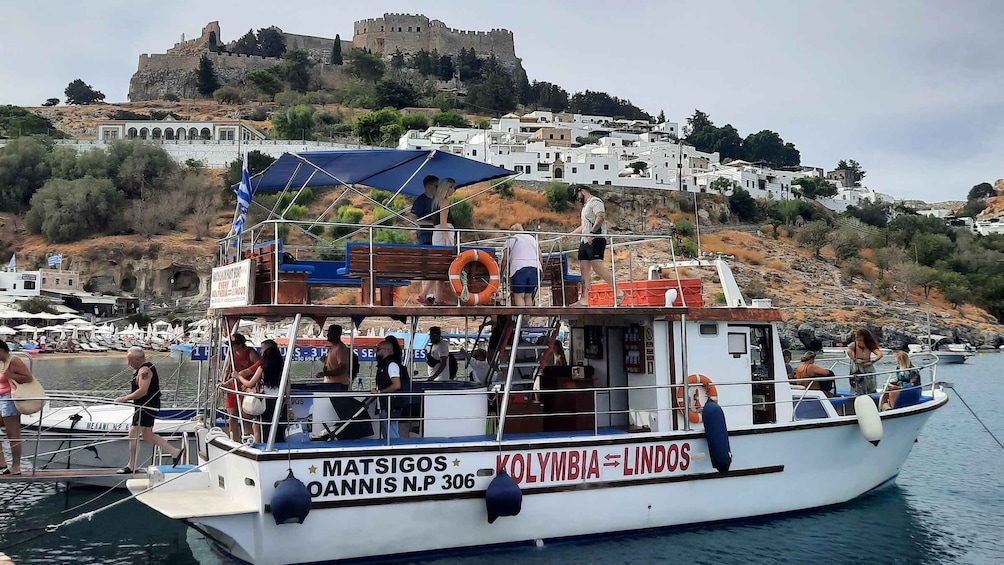A boat named 'Matsigos' docked at a harbor with a backdrop of a historic castle and white buildings. 3
