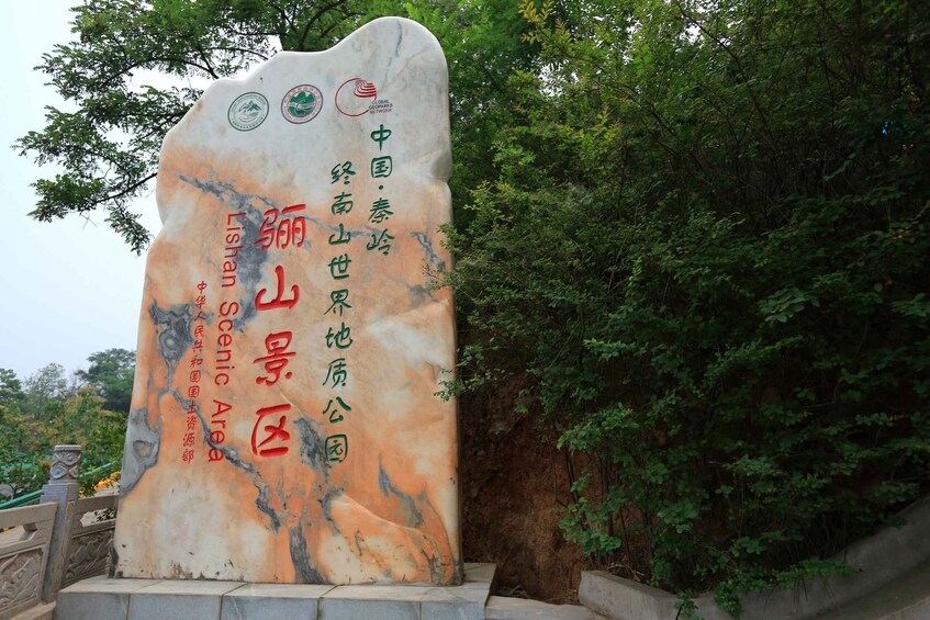 A large, vertically oriented stone monument with inscriptions and logos, surrounded by lush greenery. 4