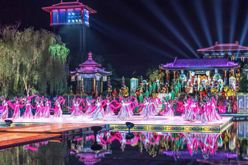 A nighttime outdoor performance with dancers in pink costumes on a stage, surrounded by traditional architecture and illuminated by spotlights. 2