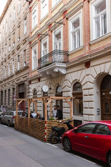 A red car parked on the street, a wooden structure with people sitting, and a building with red brick and white windows. 2