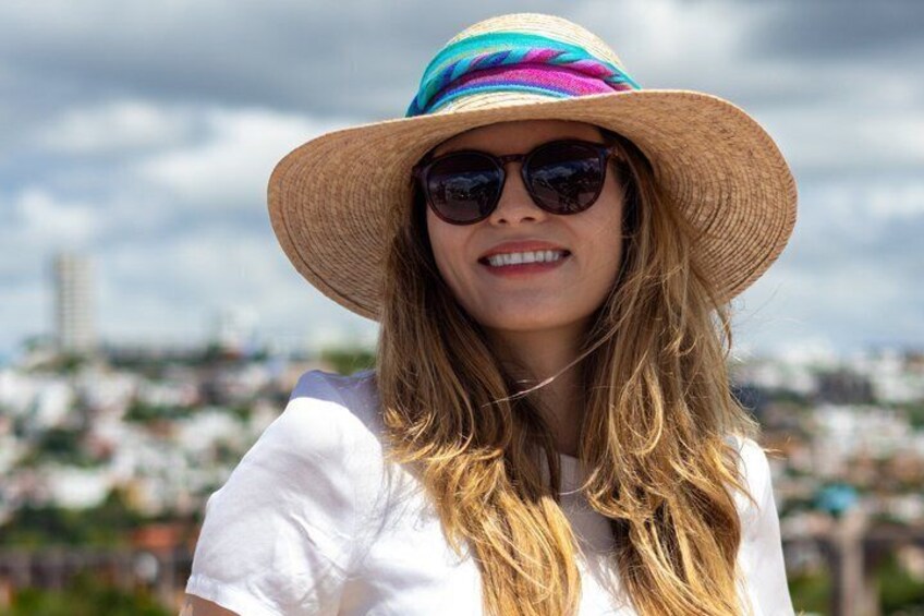 A person wearing a straw hat and sunglasses, standing outdoors with a cityscape in the background. 2