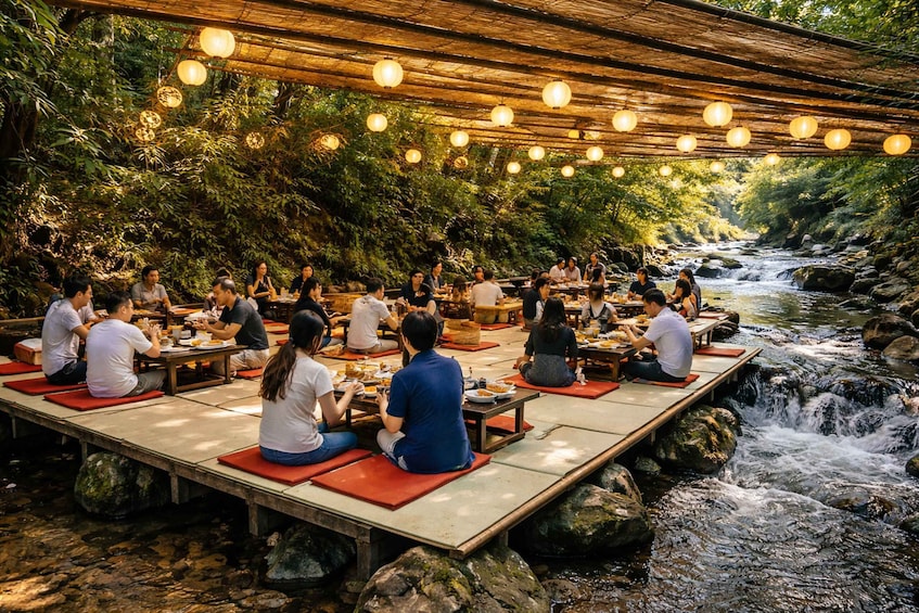 People sitting on tatami mats at tables along a riverside, under a wooden structure with hanging lights. 3