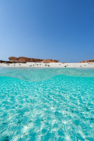 A clear turquoise sea with a sandy beach and rocky formations in the background. 4