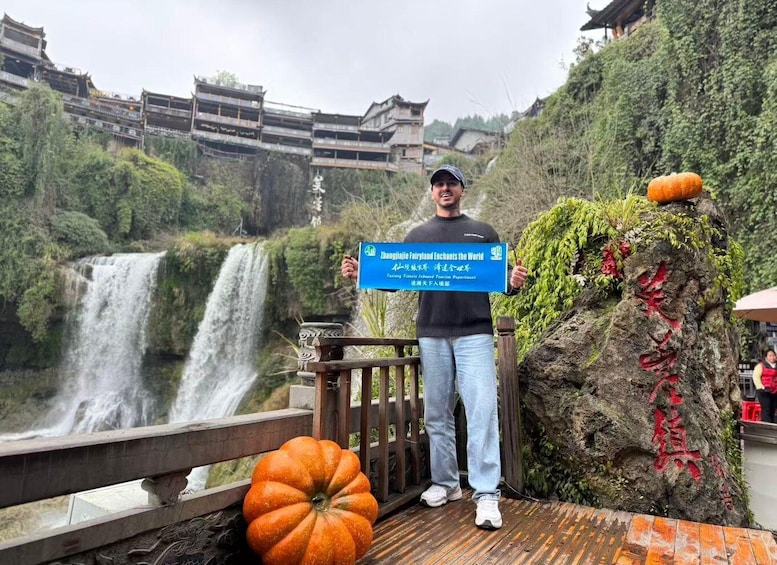 A person standing on a wooden bridge with a waterfall in the background, holding a blue sign, and a pumpkin on a rock beside them. 5