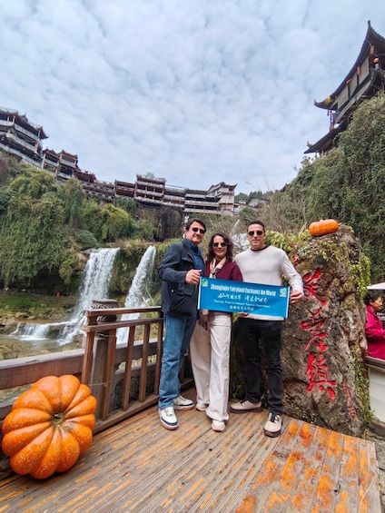 A group of people posing with a sign in front of a traditional building and a waterfall. 2