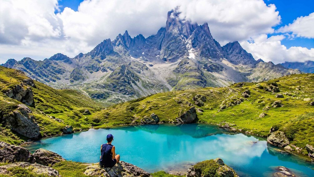 A person sitting by a turquoise lake surrounded by green hills and a mountain range. 1