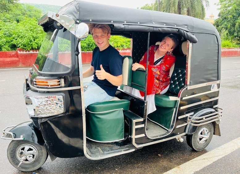 A man and a woman in a green auto-rickshaw on a road with greenery and a building in the background. 1