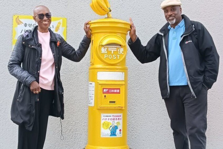 Two people standing next to a yellow post box with Japanese text. 5