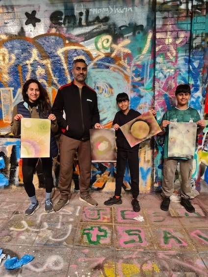 Four people holding up painted canvases in front of a graffiti-covered wall. 4
