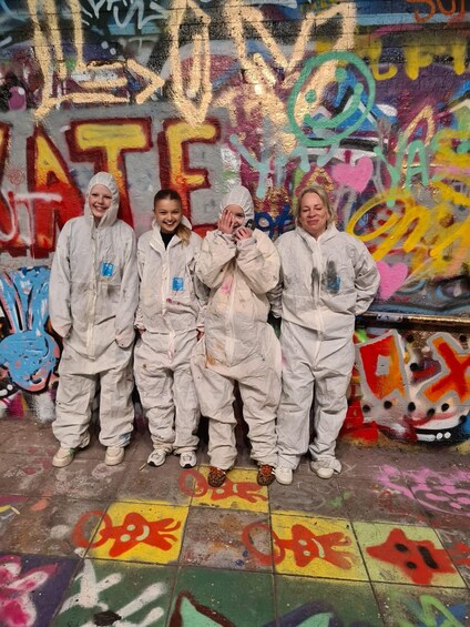Four people in white protective suits standing in front of a colorful graffiti wall. 1