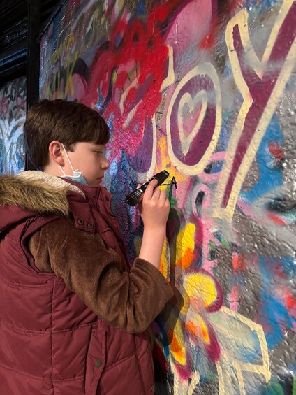 A person wearing a brown jacket with a fur collar, holding a spray paint can, standing in front of a colorful graffiti wall. 3