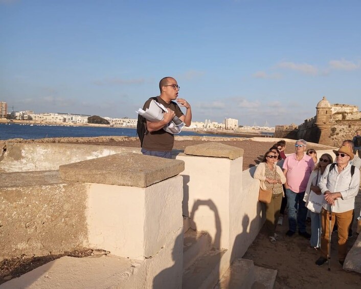 A person giving a tour, standing on a concrete platform by the sea, with a historic fort in the background. 3