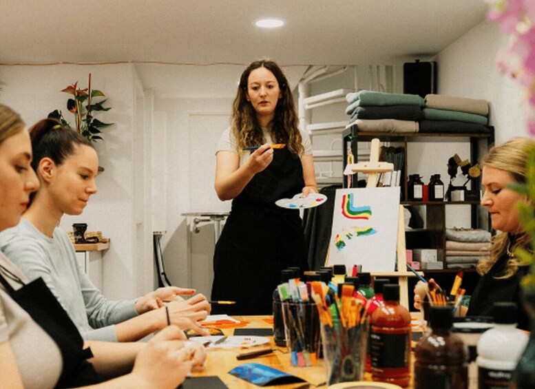 A woman painting a colorful abstract on a canvas, surrounded by art supplies and seated individuals. 1