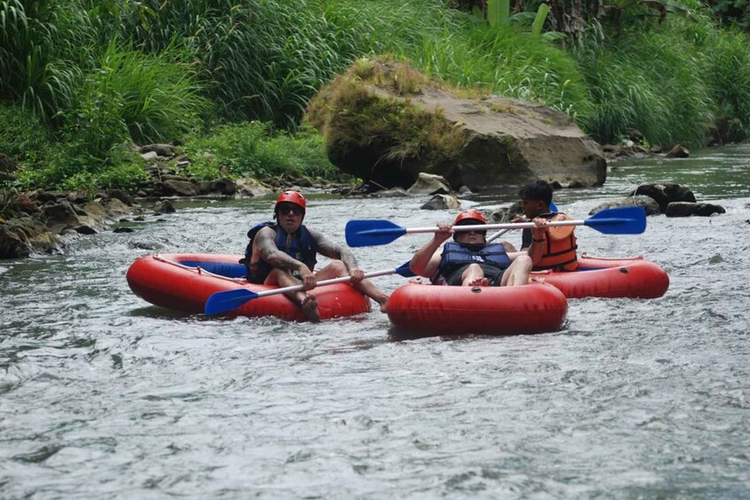 Two people in red inflatable kayaks paddling down a river surrounded by greenery. 1