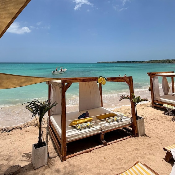 A wooden bed with cushions on a sandy beach, overlooking turquoise waters. 1