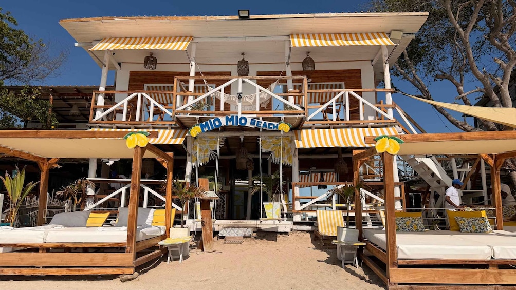 A beachside building with a sign reading 'RIO MIO BEACH' and outdoor seating with yellow cushions. 2
