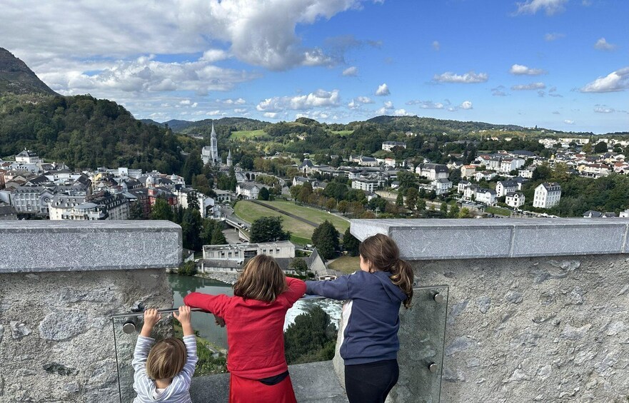 Three people overlooking a town with a prominent church spire and surrounding greenery. 1