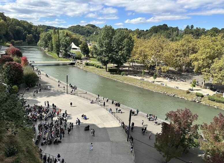 A riverside promenade with people walking and sitting, surrounded by trees and a bridge. 2