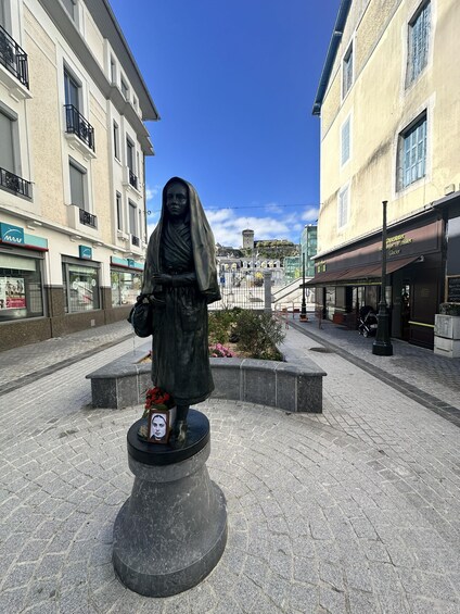 A statue of a woman on a pedestal with flowers, set in a plaza with buildings and a clear sky. 3