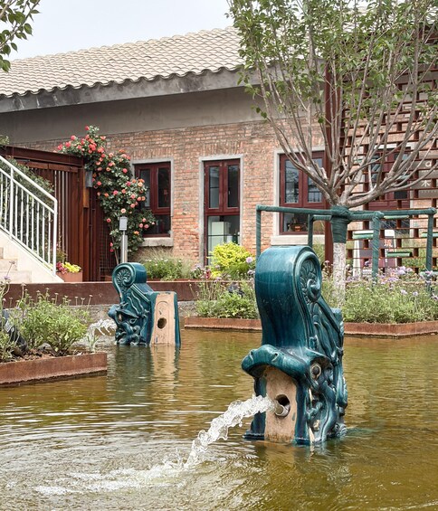 A fountain with water spouting from its center, surrounded by a garden with various plants and flowers, set against a brick building with a tiled roof. 4