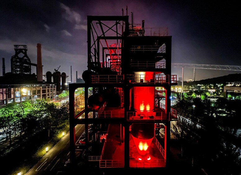 A large industrial facility with multiple red-lit structures at night, surrounded by a cityscape and a bridge in the background. 1