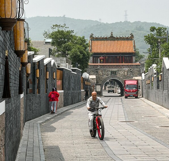 A person riding a bicycle on a paved path, with traditional architecture and a red vehicle in the background. 2