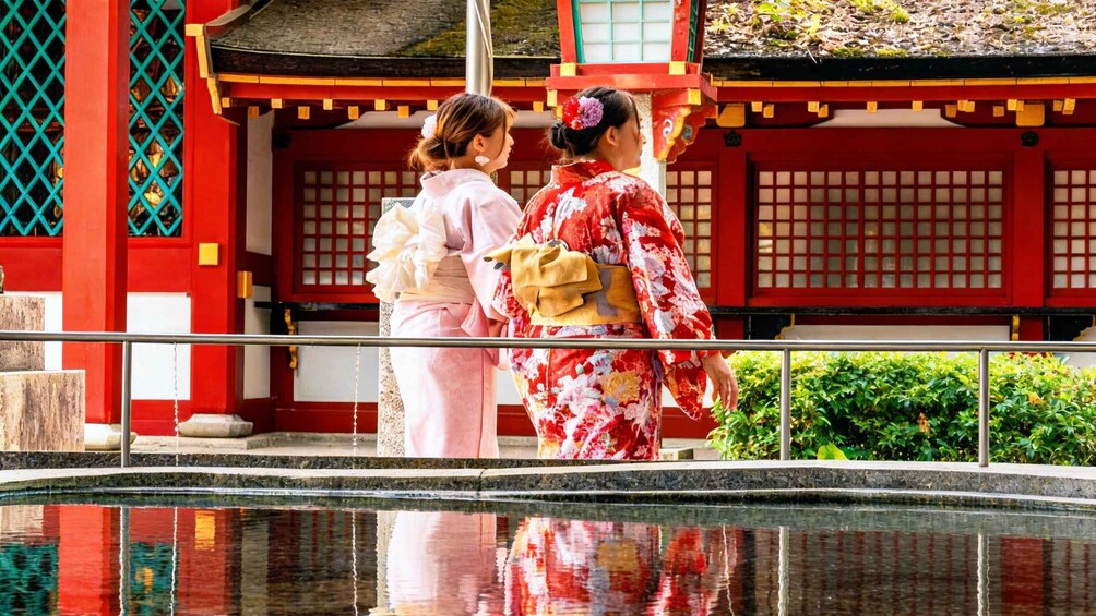 Two people in traditional Japanese attire standing by a reflective water feature in front of a red building with a tiled roof. 3