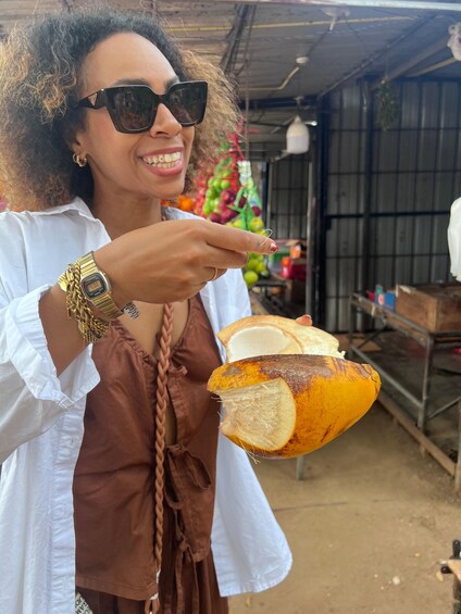 A person holding a coconut with a visible hole, wearing a white shirt and a brown dress, with a market stall in the background. 5