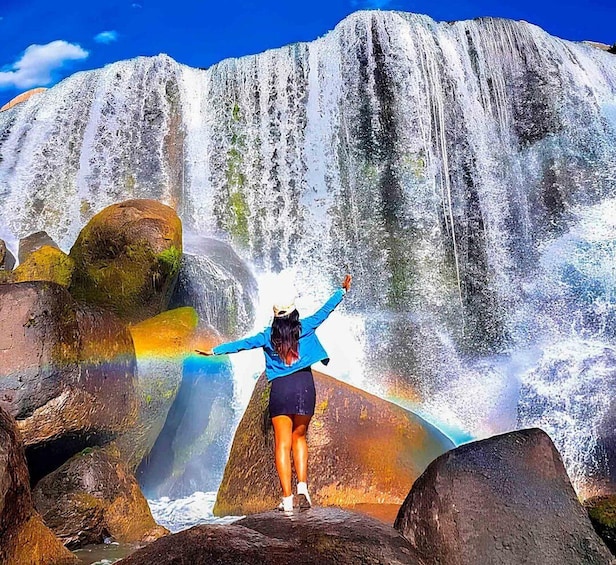 A person standing on rocks with arms raised, facing a large waterfall. 1