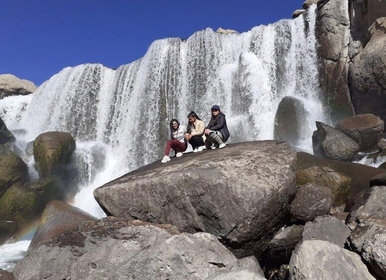 A waterfall cascading over rocks with three people sitting on a large boulder in front. 2