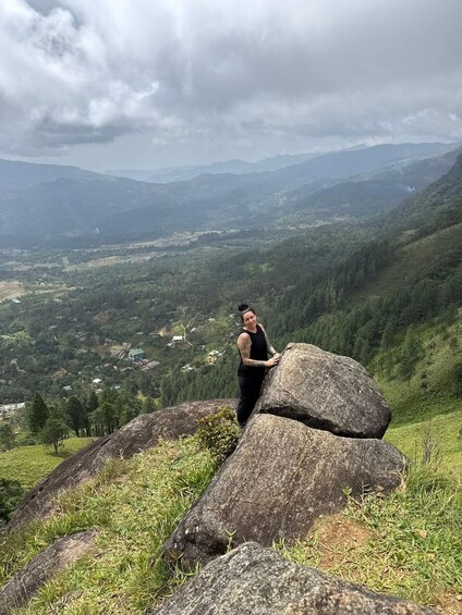 A person standing on a grassy hill with large boulders, overlooking a valley with scattered houses and dense forests under a cloudy sky. 1