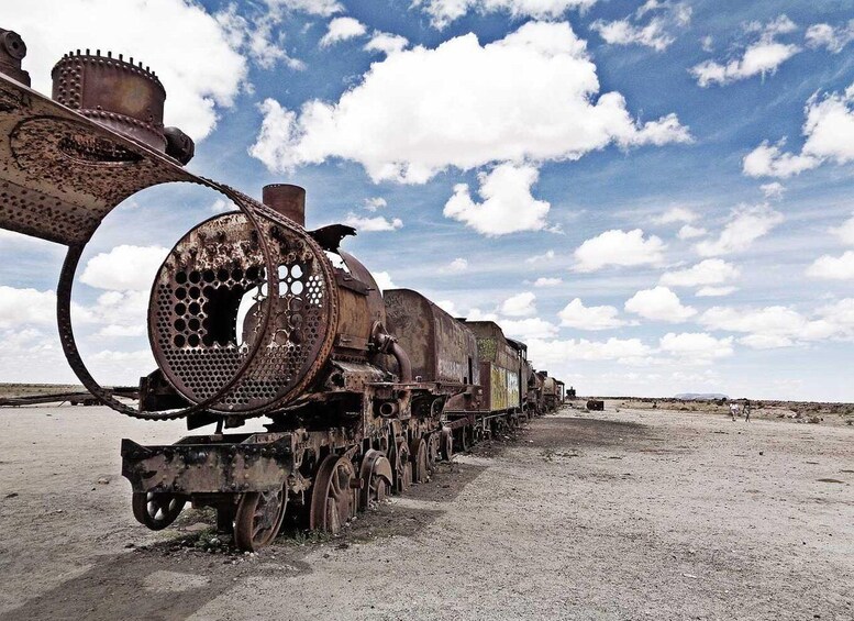 A rusted, abandoned train with a large cylindrical engine and multiple carriages, parked in a barren landscape with a clear sky. 5