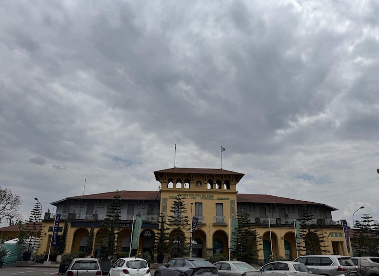 A large yellow building with arched windows and a central tower, surrounded by parked cars and trees. 1