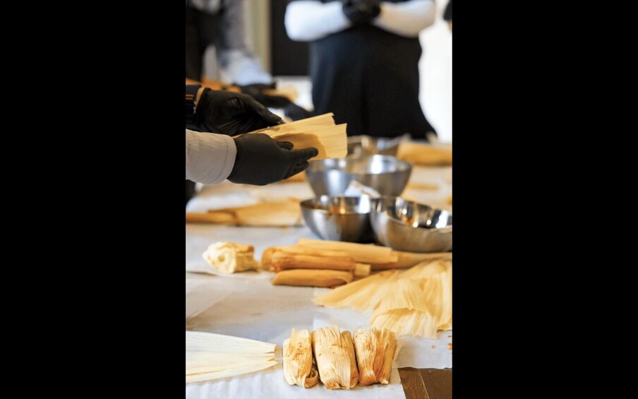 A person preparing tamales with masa and filling, surrounded by bowls and wooden sticks on a table. 1