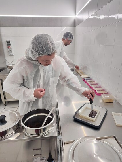 Two chefs in a kitchen preparing food, wearing hairnets and gloves, with stainless steel equipment and containers of ingredients. 2