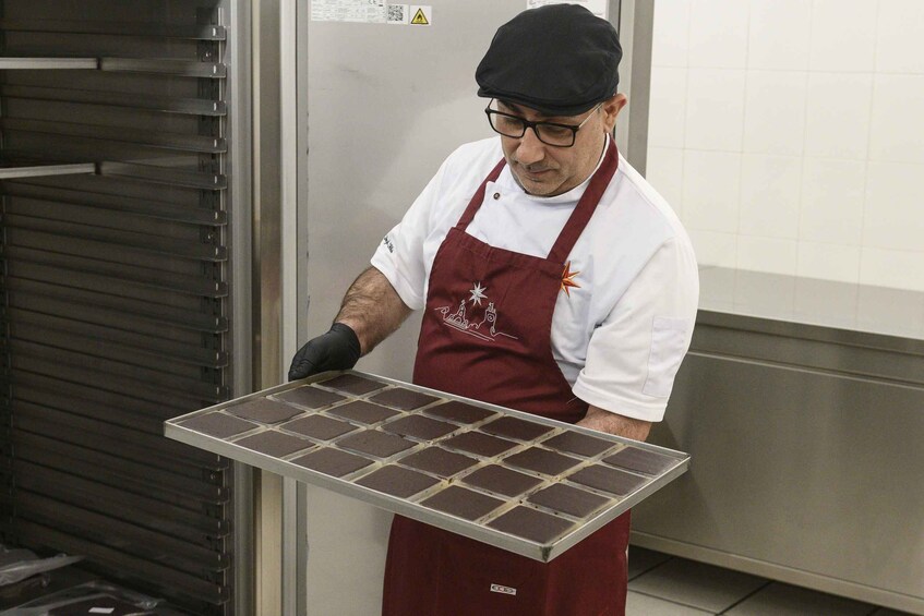 A person wearing a white shirt and a red apron with a logo, holding a tray with square items. 1