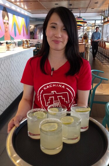 A person in a red shirt with 'KACHINA' text, holding a tray with drinks and a lemon wedge. 3