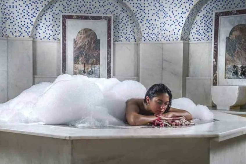 A person in a bathtub with white bubbles, surrounded by marble walls and framed artwork. 3