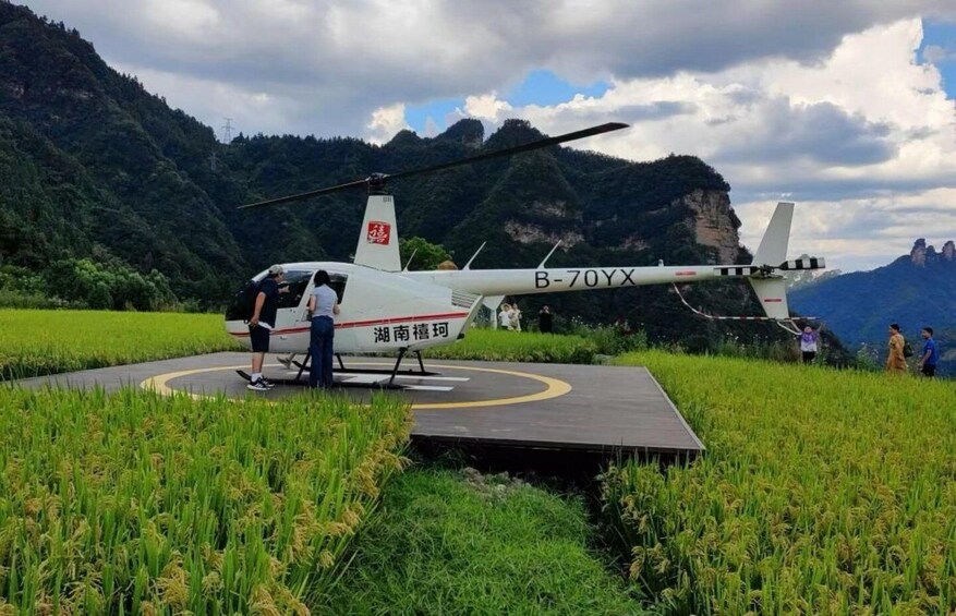 A helicopter with registration B-70YX on a landing pad surrounded by rice fields and mountains in the background. 2