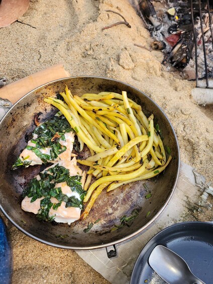 A pan with grilled fish, French fries, and greens on a sandy surface. 2