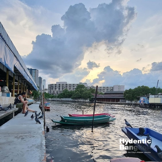 A waterfront scene with a woman and child by a building, boats on the water, and urban buildings in the background. 1