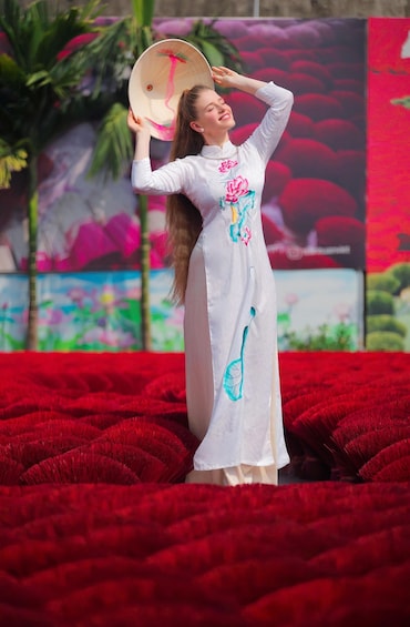 A person in a white dress with floral embroidery standing on a red carpet, holding a hat. 2