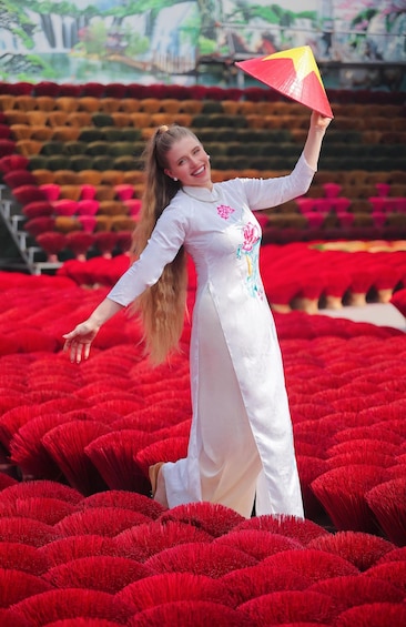 A person in a white dress with floral embroidery, holding a red and yellow fan, standing in a field of red flowers. 5