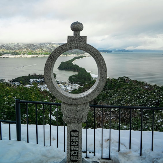 A stone monument with a circular mirror reflecting a coastal landscape, surrounded by snow and a metal fence. 5