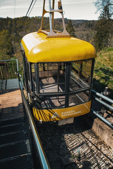 A yellow cable car with 'SIGULDA ADVENTURES' written on it, suspended by cables, with a forested landscape in the background. 4