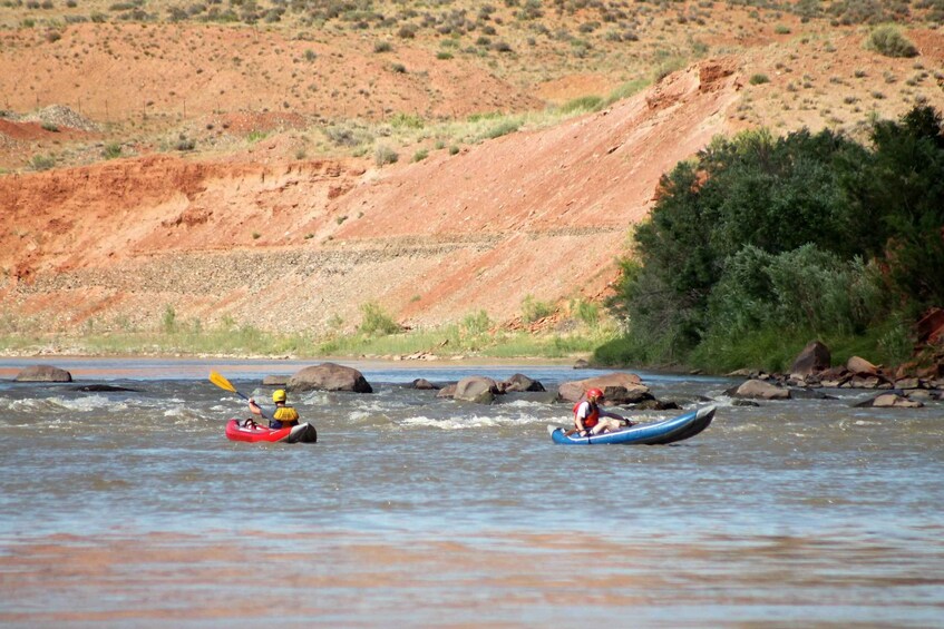 Two kayakers on a river with a rocky riverbed and sparse vegetation in the background. 5