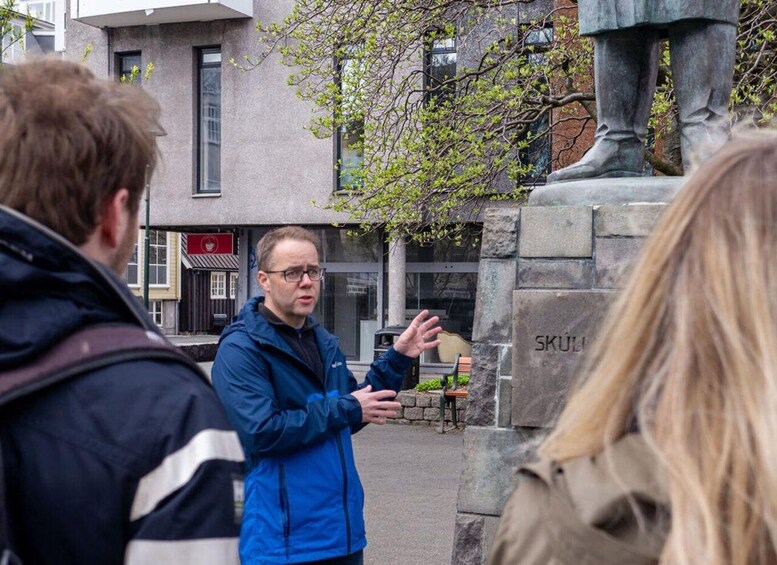 A person in a blue jacket gesturing, standing in front of a stone monument with 'SKÚL' inscribed, in an urban setting with buildings and a tree. 1