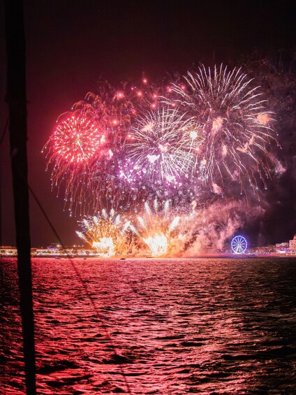 A night scene with fireworks over water, a Ferris wheel, and a distant shoreline. 1